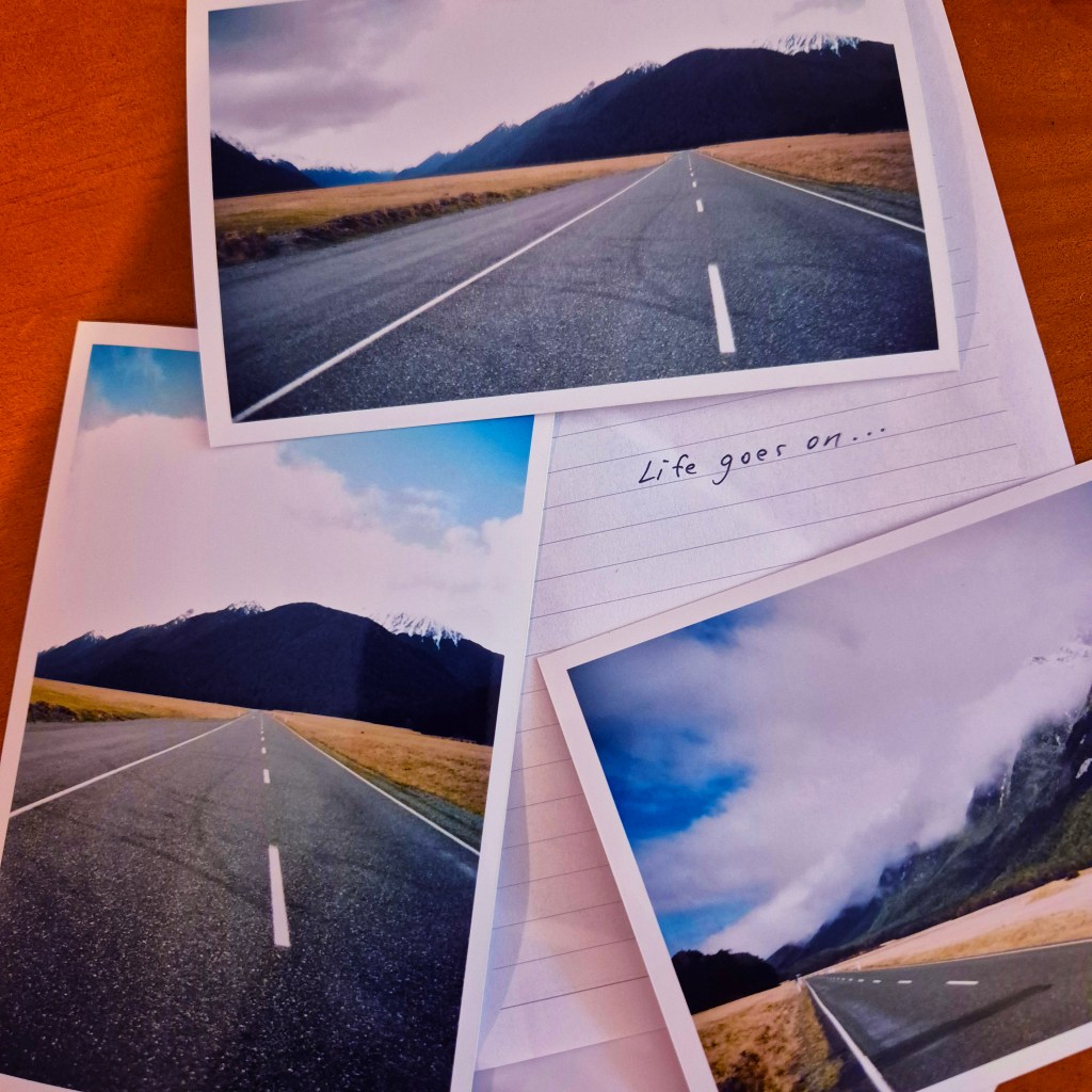 Three landscape photographs of a winding road surrounded by mountains and grasslands, displayed on a wooden surface, with a handwritten note that says 'Life goes on...'.